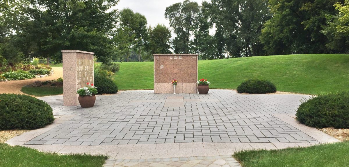Two columbarium walls face the center of a cross shaped pad of pavers accented with a shrub in each corner.