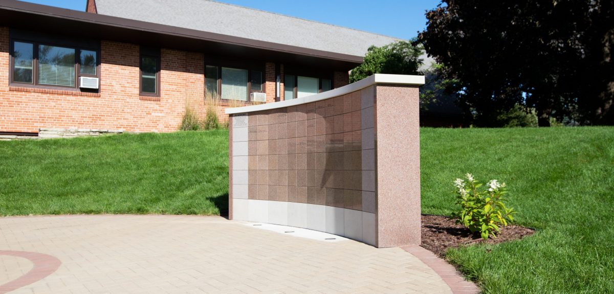 At the bottom of a grassy hill a columbarium wall sits on a circular pad of pavers.