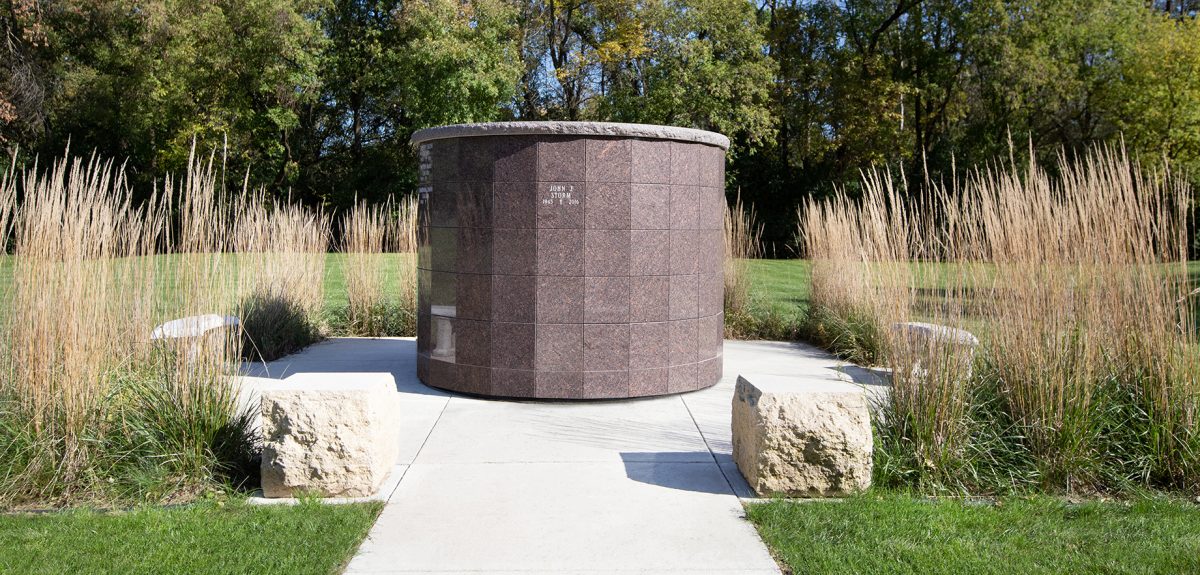 A cement sidewalk leads to a Pre-Assembled columbarium and is adorned with two rough cut stone blocks.