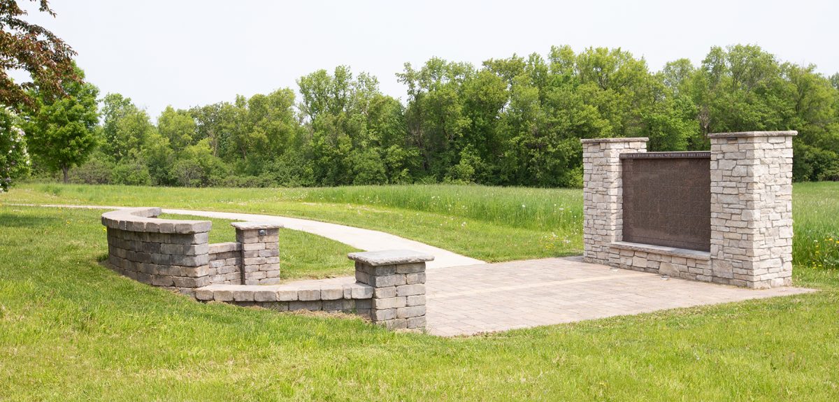 A retaining wall, a pad of paving stones, and a rectangular columbarium define a memorial space nestled in nature.