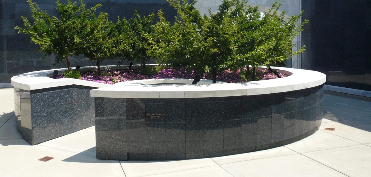 Two towering columbarium walls stand behind a shorter circular columbarium structure adorned with small, flourishing trees and light pink flowers.