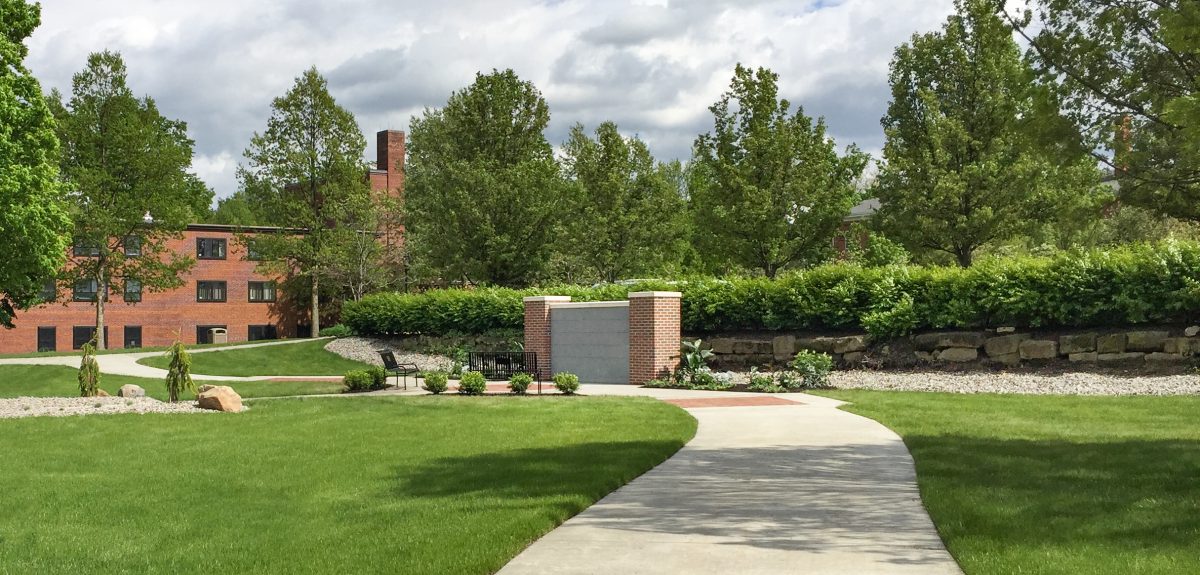A curved sidewalk leads to a memorial space with benches, attractive plantings, and a columbarium wall on campus.
