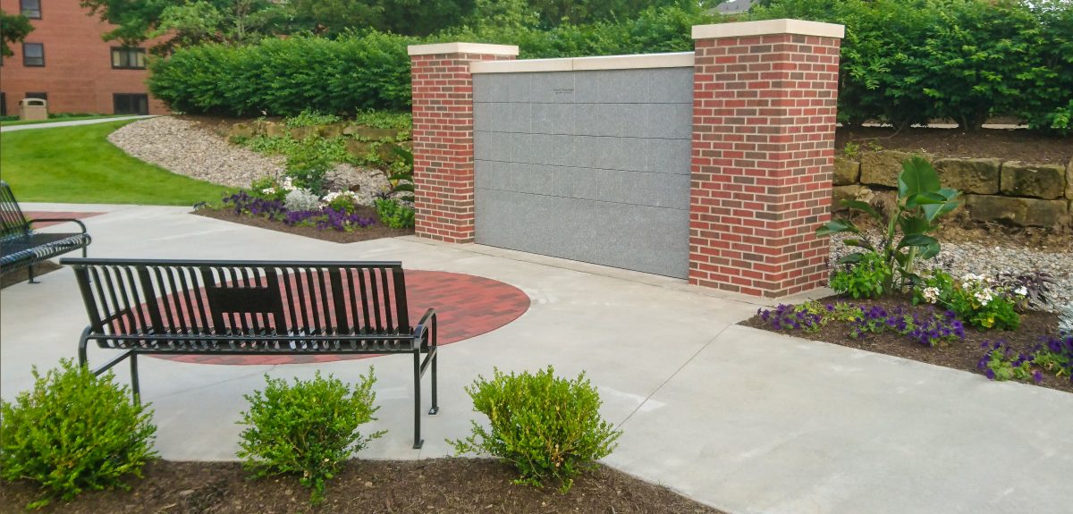 Well manicured landscaping surrounds a columbarium wall with two metal benches.