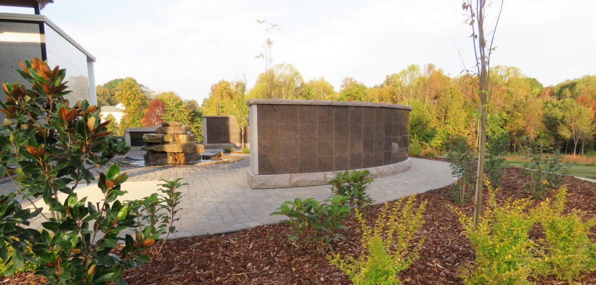 A rough cut stone fountain reflects bright slivers of sunlight cast over two curved columbaria walls.