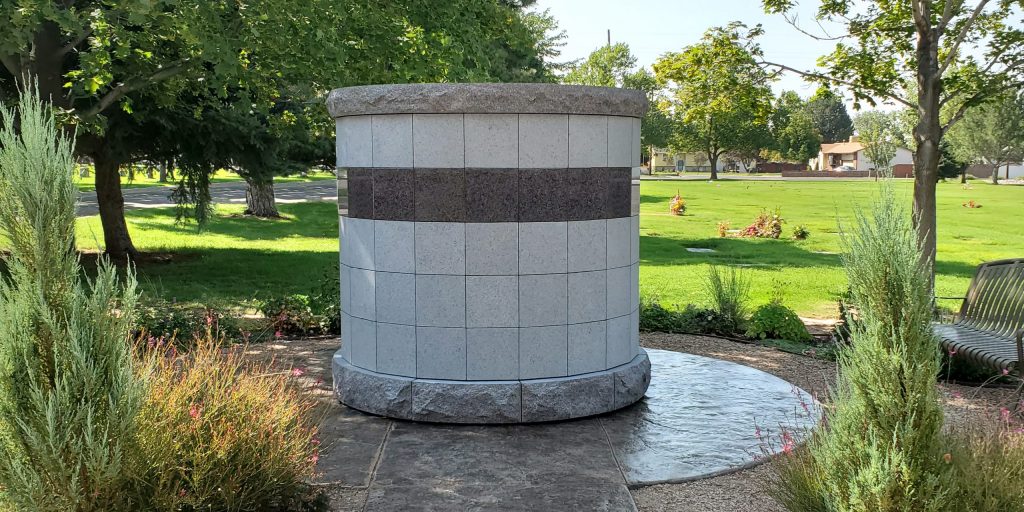 A Canterbury Ossuarium surrounded by attractive plantings and two metal benches sits atop a textured concrete pad in a cemetery.